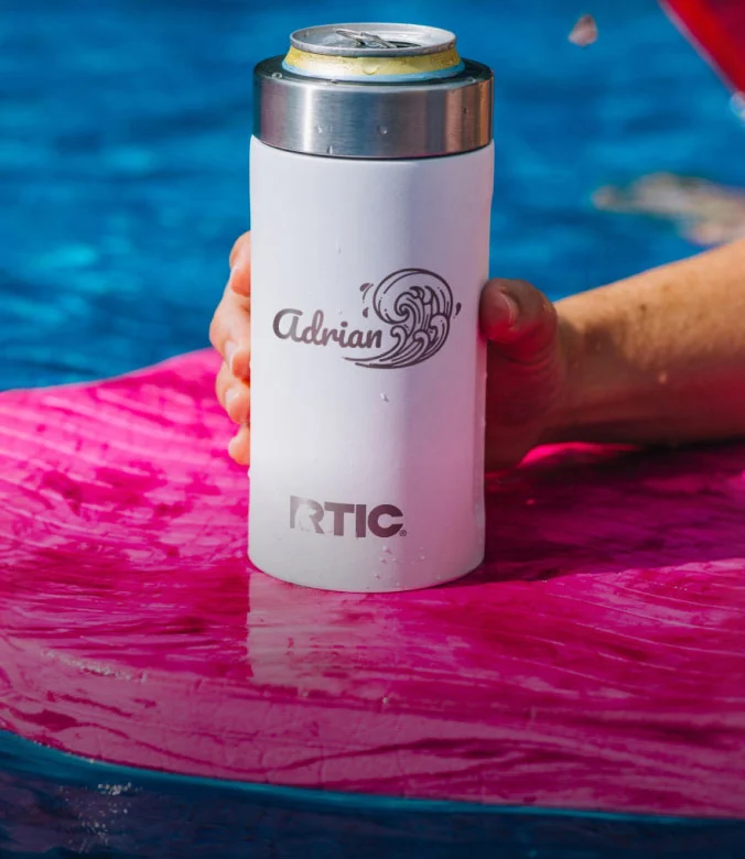White RTIC insulated can cooler with a personalized name and wave design, holding a beverage by a swimming pool.